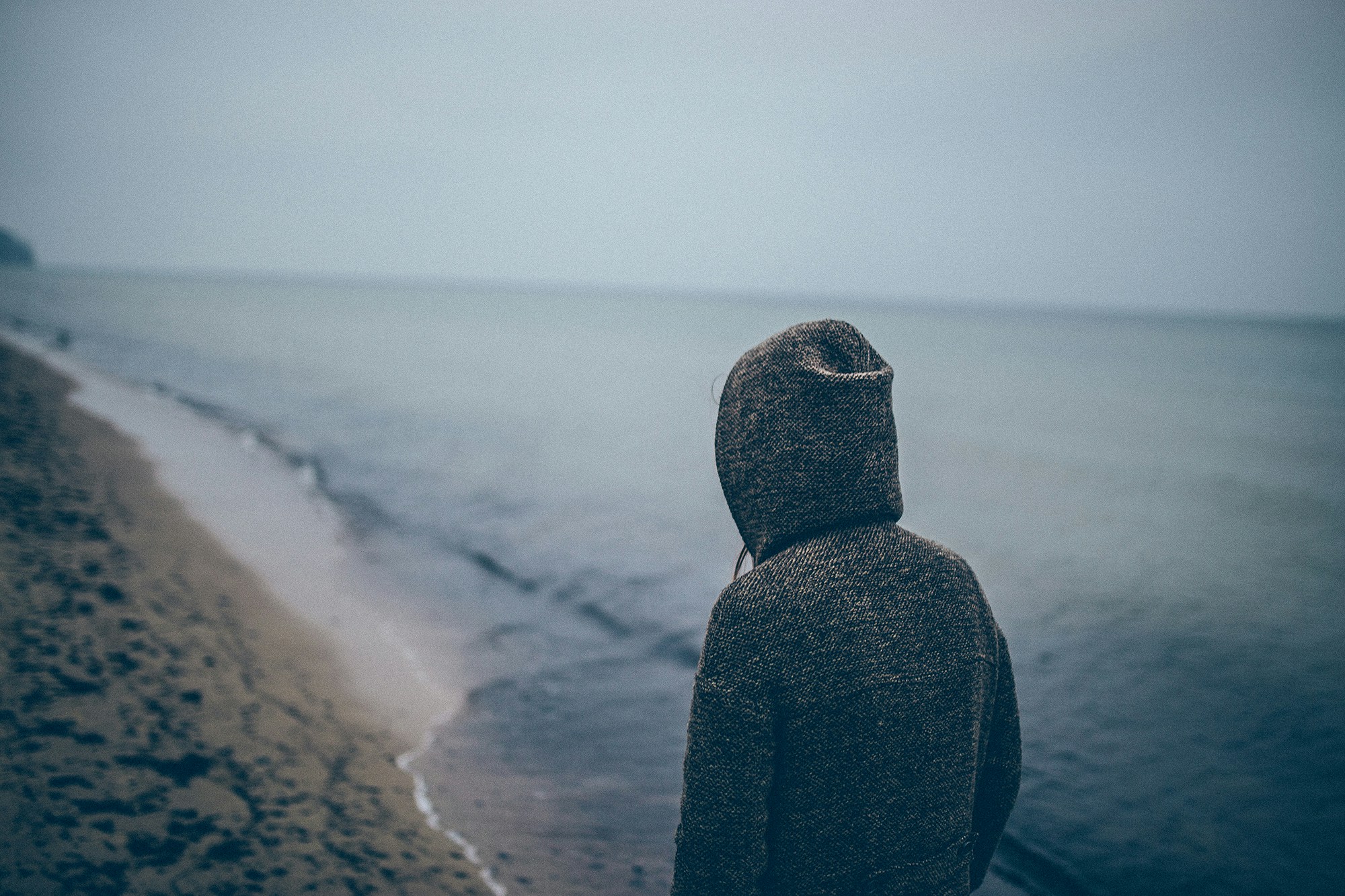 hooded figure walking away from the photographer along a  grey lonely beach.